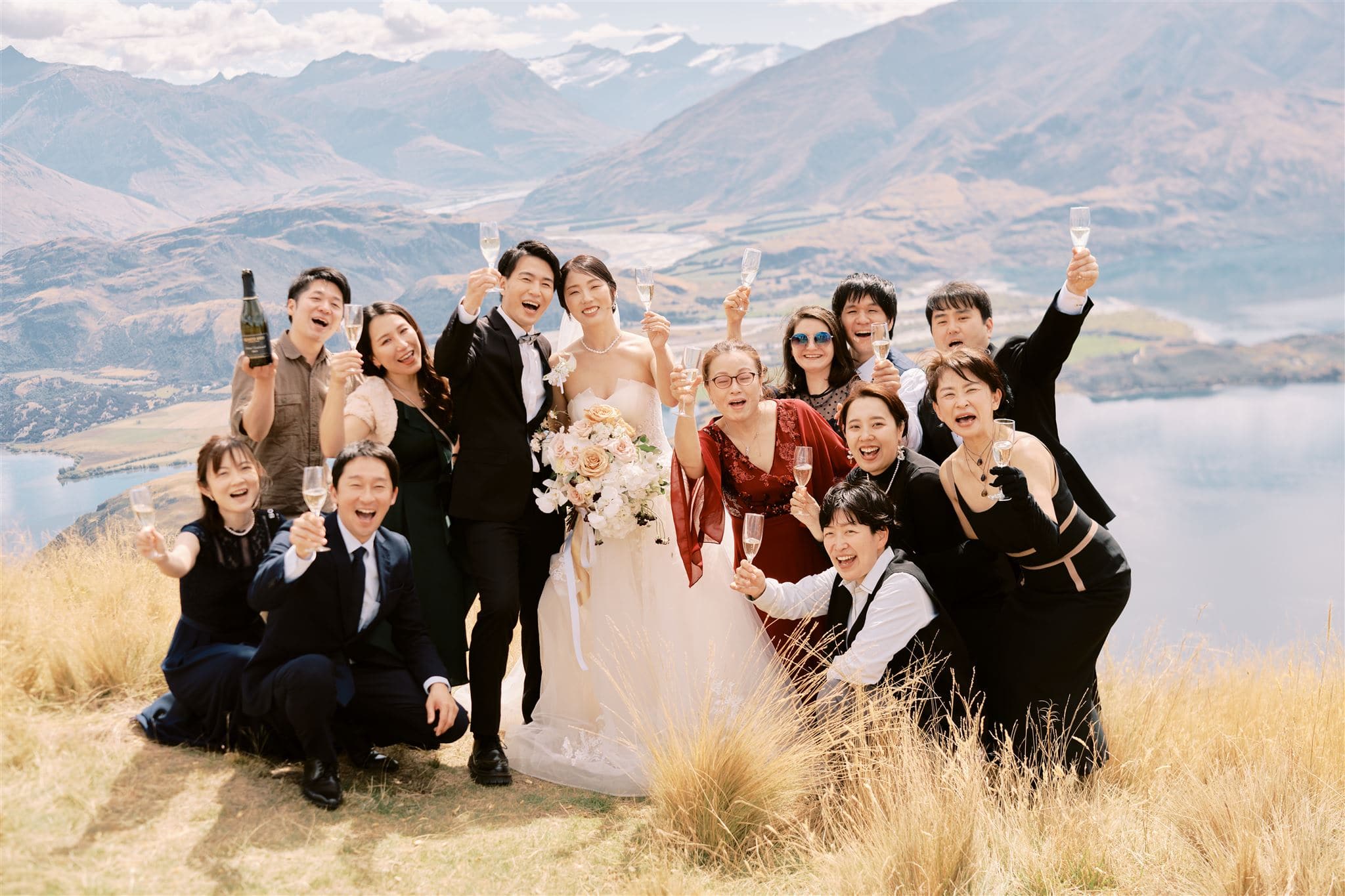 Queenstown New Zealand Heli Wedding Elopement Photographer クイーンズタウン　ニュージーランド　エロープメント 結婚式 | A wedding group celebrating outdoors with a mountainous landscape and a lake in the background, captured by YURI, a Queenstown Wedding Photographer.